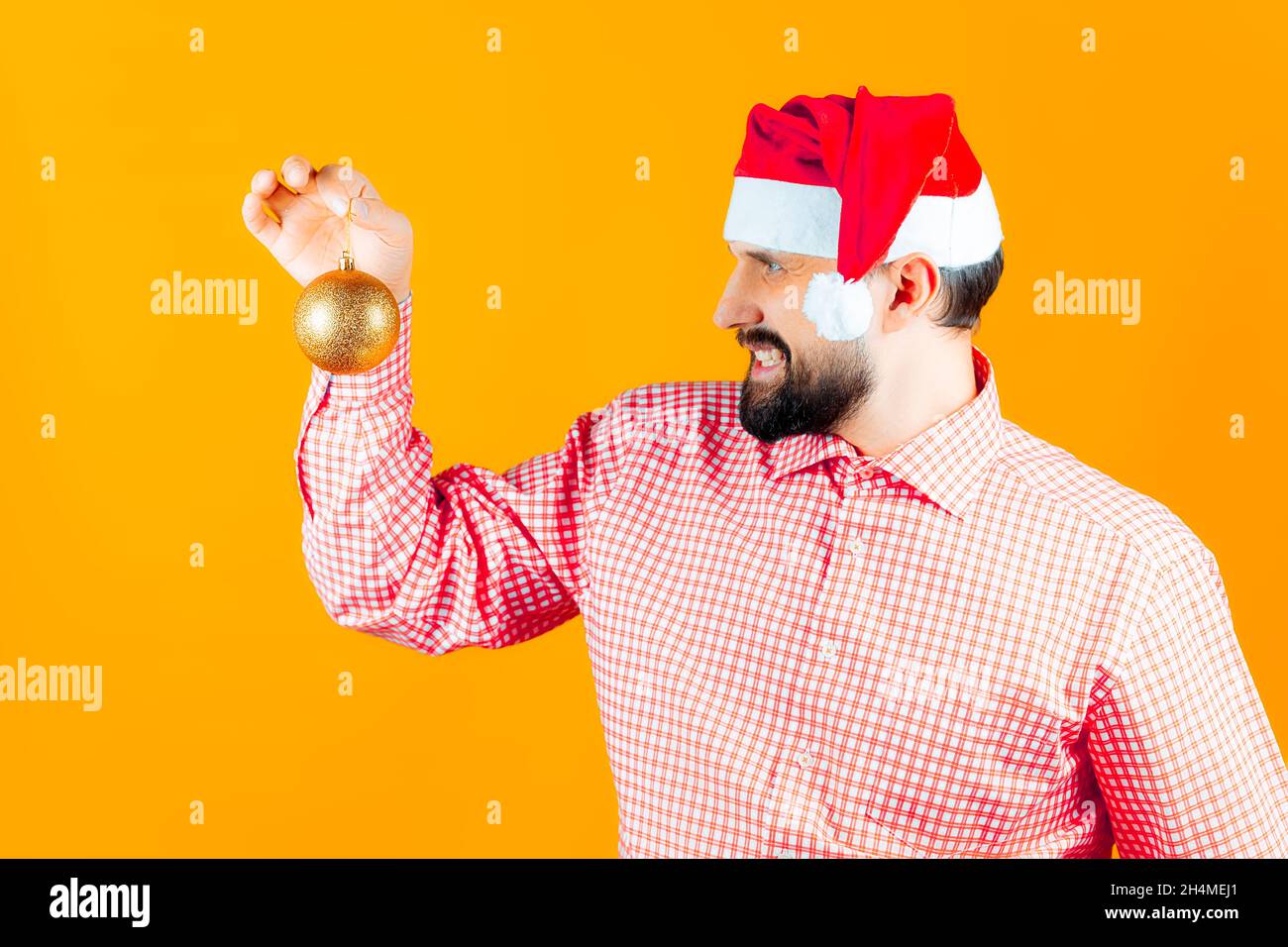 Un homme dans les chapeaux du nouvel an du Père Noël et une chemise à carreaux rouges examine attentivement les jouets du nouvel an - une balle de couleur or. Banque D'Images