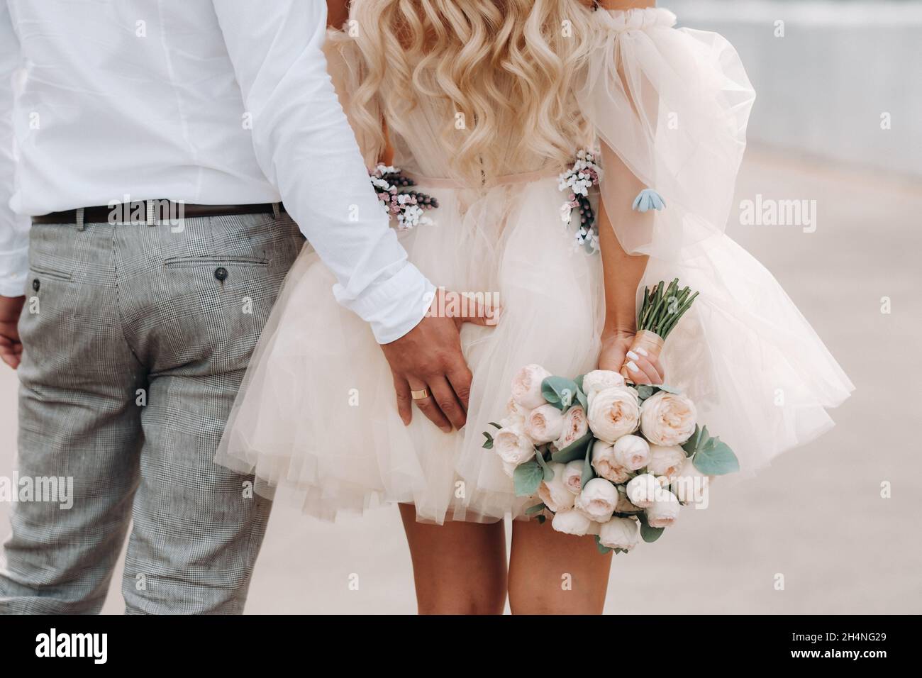 Le marié met sa main derrière sa mariée.La mariée dans une robe courte tient un bouquet dans ses mains de derrière. Banque D'Images