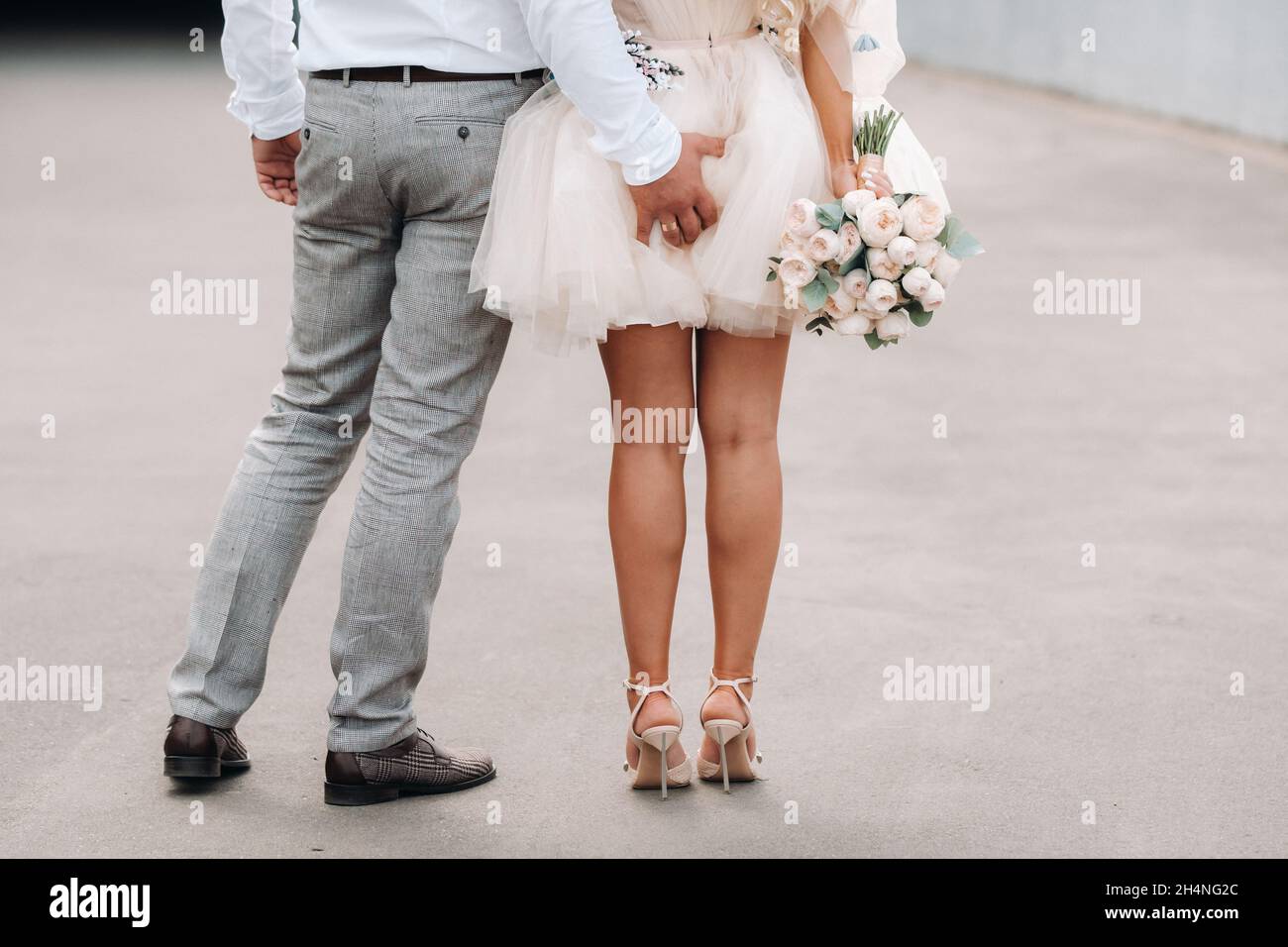 Le marié met sa main derrière sa mariée.La mariée dans une robe courte tient un bouquet dans ses mains de derrière. Banque D'Images