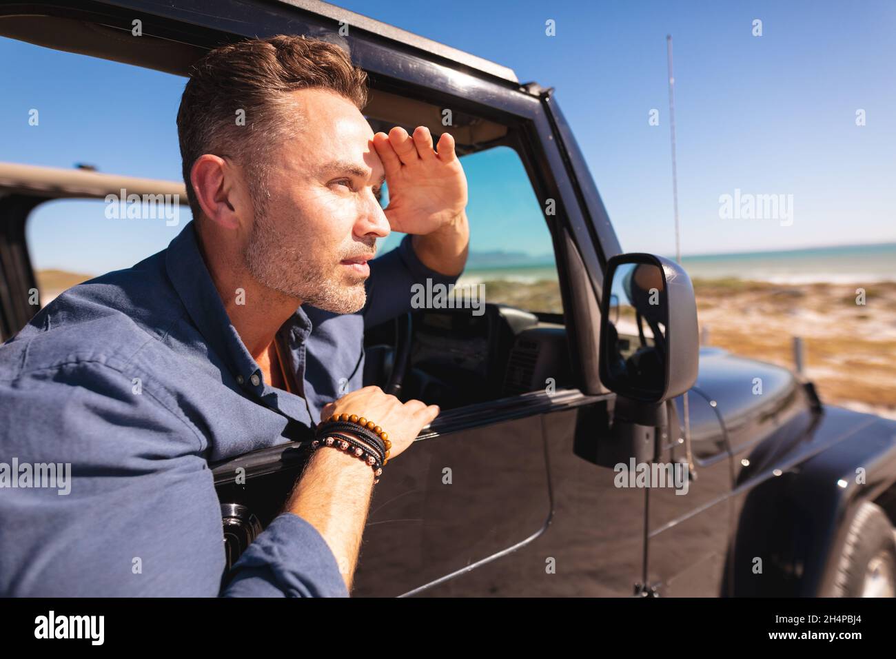Homme caucasien attentionné assis en voiture au bord de la mer en protégeant les yeux du soleil et en admirant la vue Banque D'Images