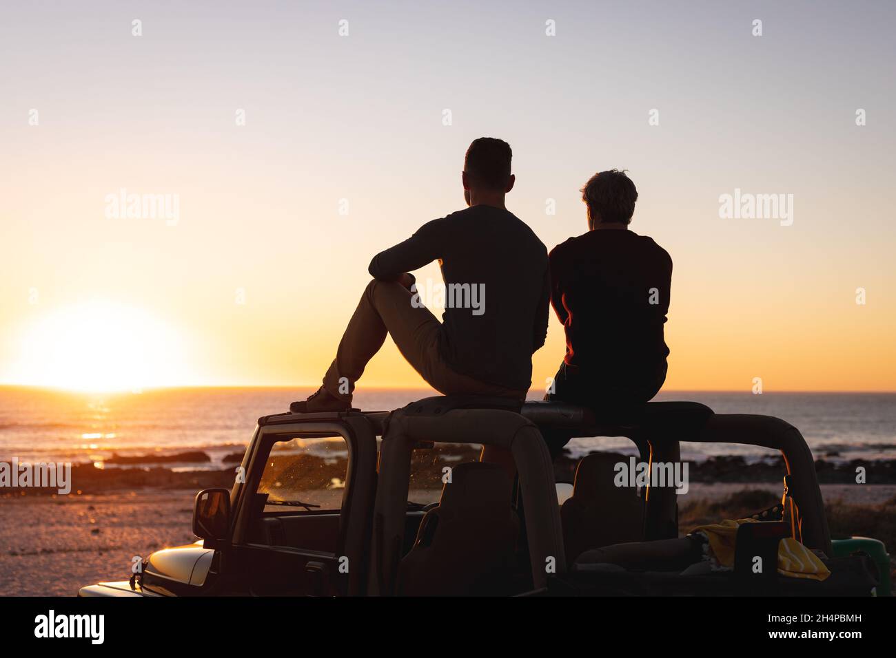 Vue arrière d'un couple de hommes gais caucasiens assis sur le toit de la voiture au coucher du soleil au bord de la mer Banque D'Images