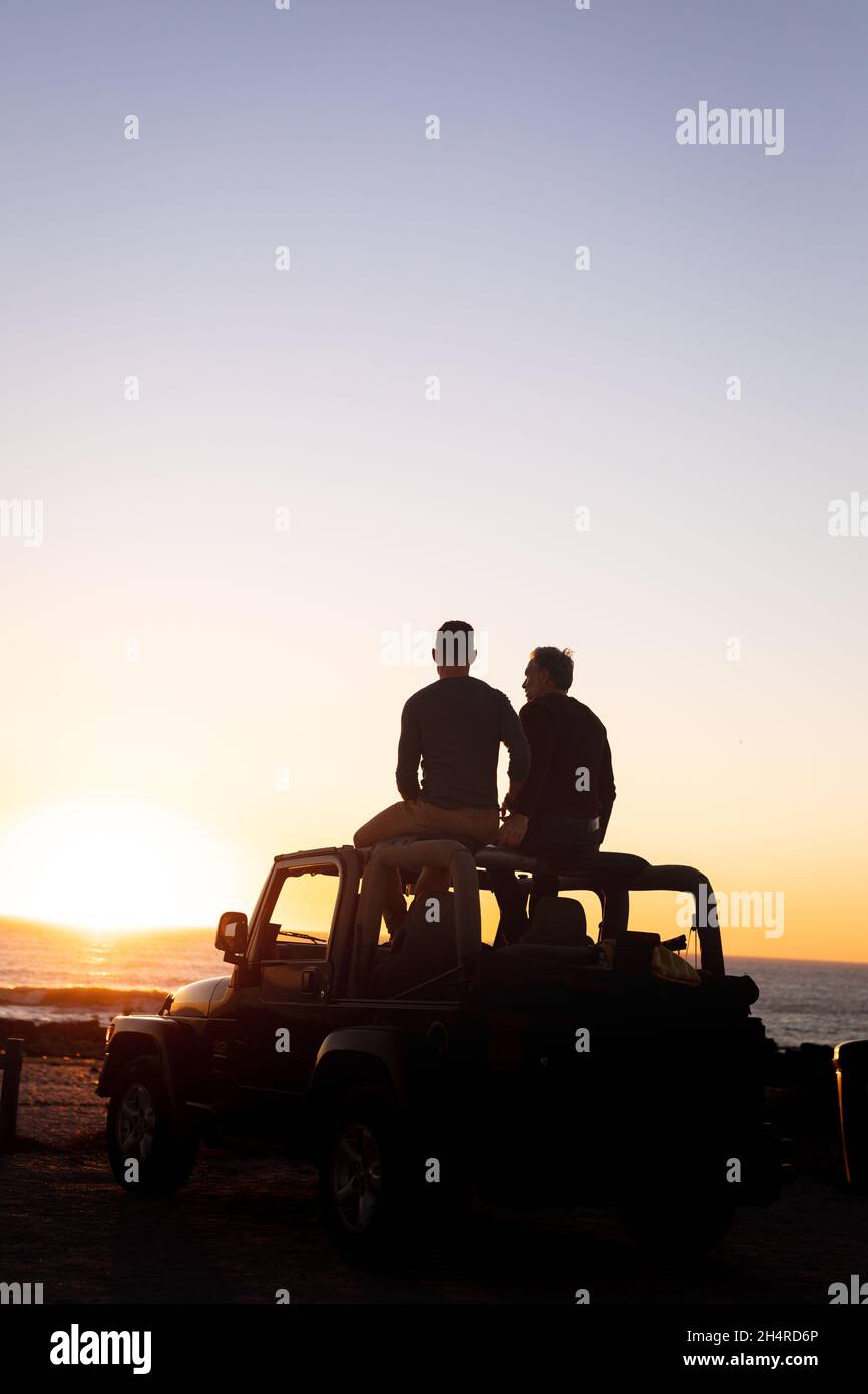 Vue arrière d'un couple de hommes gais caucasiens assis sur le toit de la voiture au coucher du soleil au bord de la mer Banque D'Images