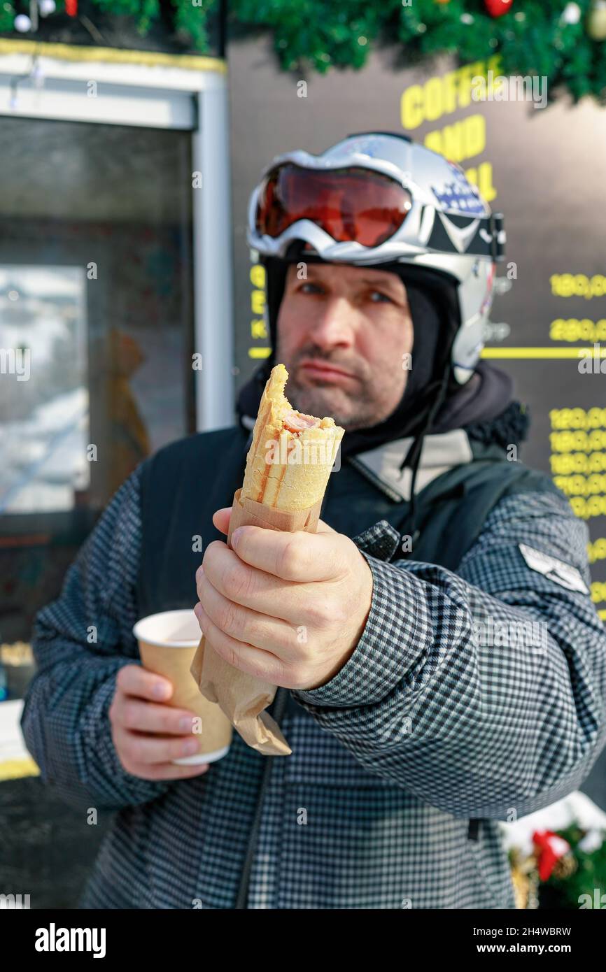 Portrait d'un snowboardeur avec un hot dog.Un homme d'origine caucasienne d'âge moyen dans un casque de sécurité sur une piste de ski se repose et boit du café chaud W. Banque D'Images