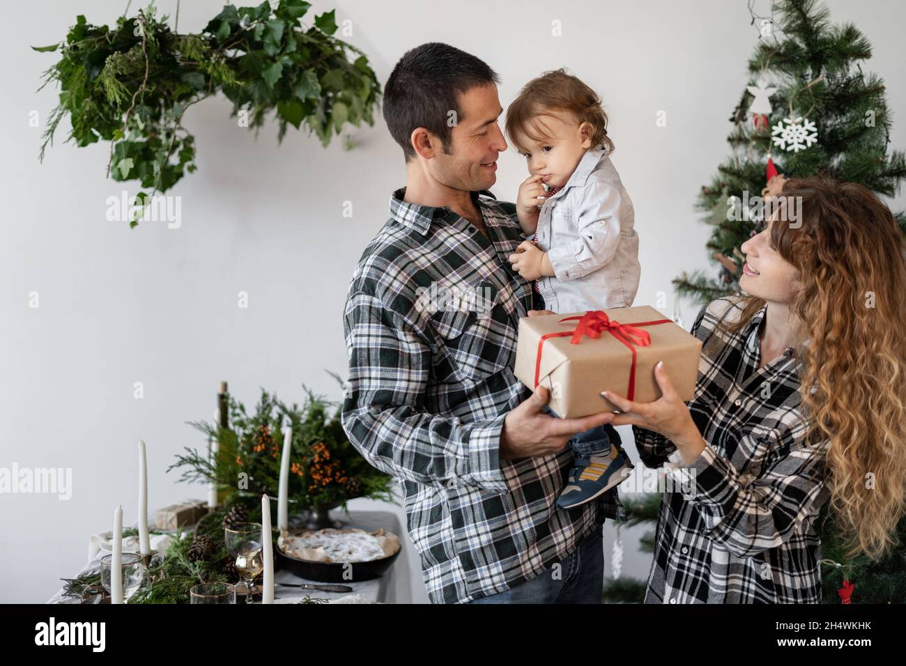 Noël à la maison, une maman de famille donne un cadeau à un petit fils assis dans les bras de son père dans le salon décoré d'un arbre de Noël et d'une table Banque D'Images