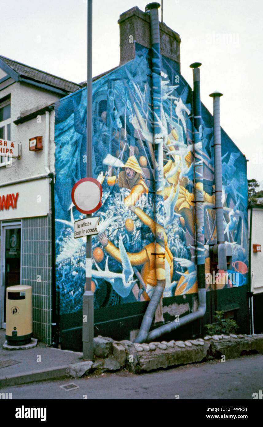 «Fishing», une fresque d’Edward Povey sur le côté d’un magasin de poissons et de puces situé au 27-29 High St, Menai Bridge Anglesey, pays de Galles, en 1987.La peinture murale a été commandée par le propriétaire de l’atelier de l’époque, M. Radford (senior), et a été peinte à base d’huile sur le rendu de ciment apprêté en juin 1980.Il représente un chalutier de pêche et des pêcheurs en mer.Il est toujours en place, mais il est défraîchi et endommagé par le temps – le bâtiment est maintenant un restaurant indien.Artiste anglais, Edward Povey (n.1951), peint 25 peintures murales sur des bâtiments du pays de Galles et du nord-ouest de l'Angleterre entre 1975 et 1985. Banque D'Images