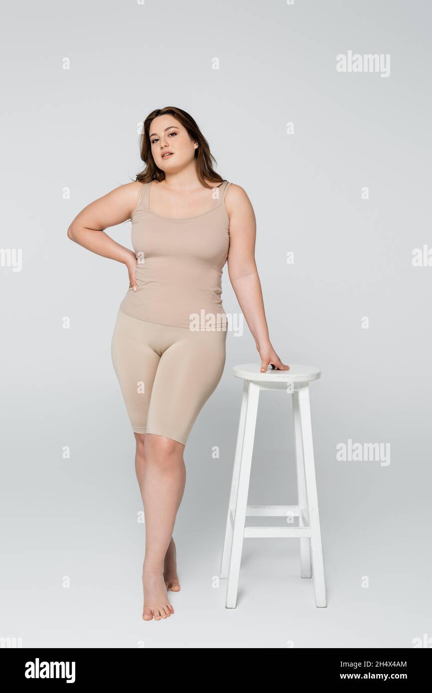Pleine longueur de jolie femme de taille plus tenant la main sur la hanche  près de la chaise sur fond gris Photo Stock - Alamy