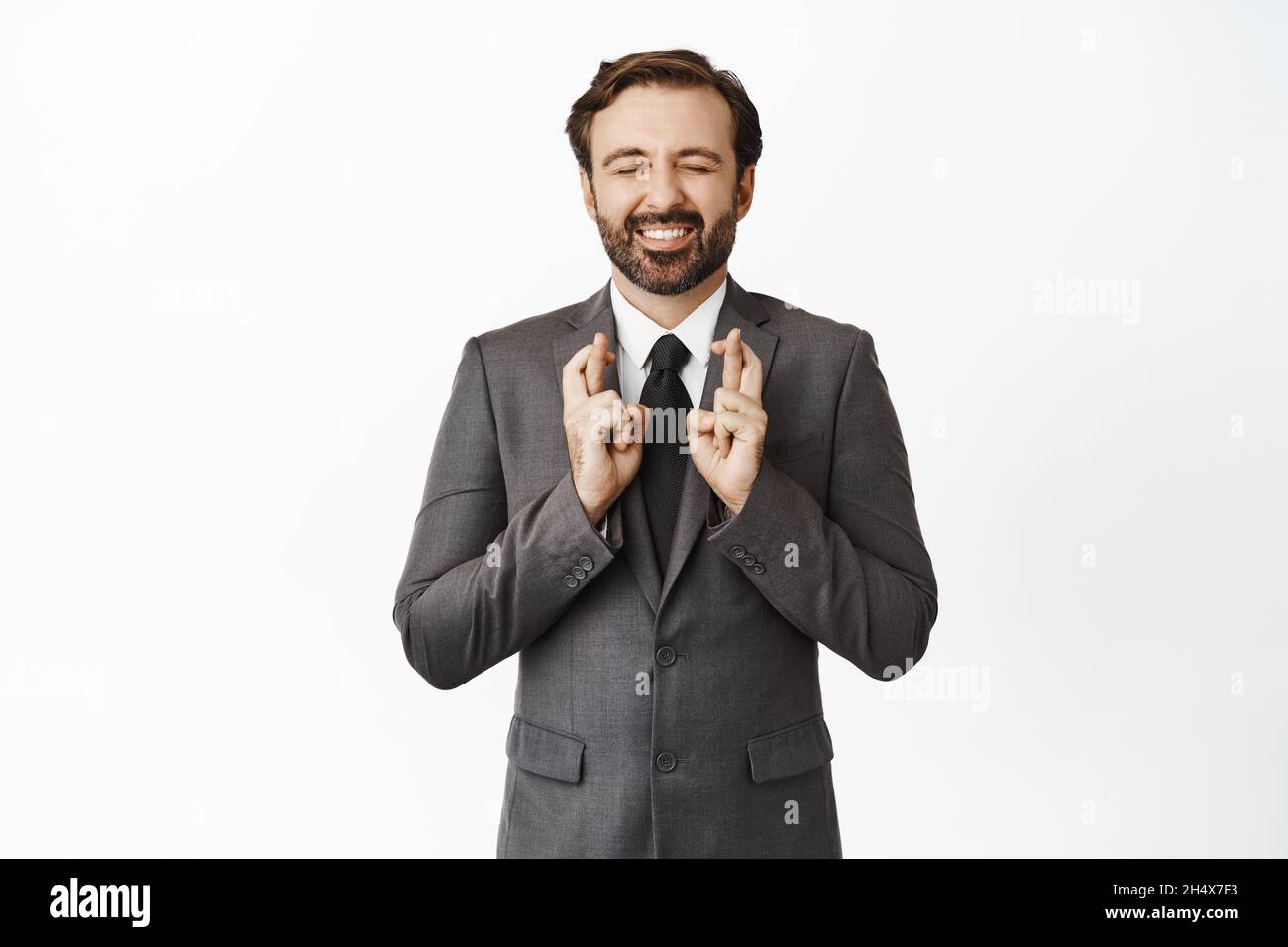 Un homme d'affaires enthousiaste croiser les doigts et les yeux, souriant tout en faisant le souhait, anticipant quelque chose, aspirant à obtenir un emploi, se tenant sur le blanc Banque D'Images