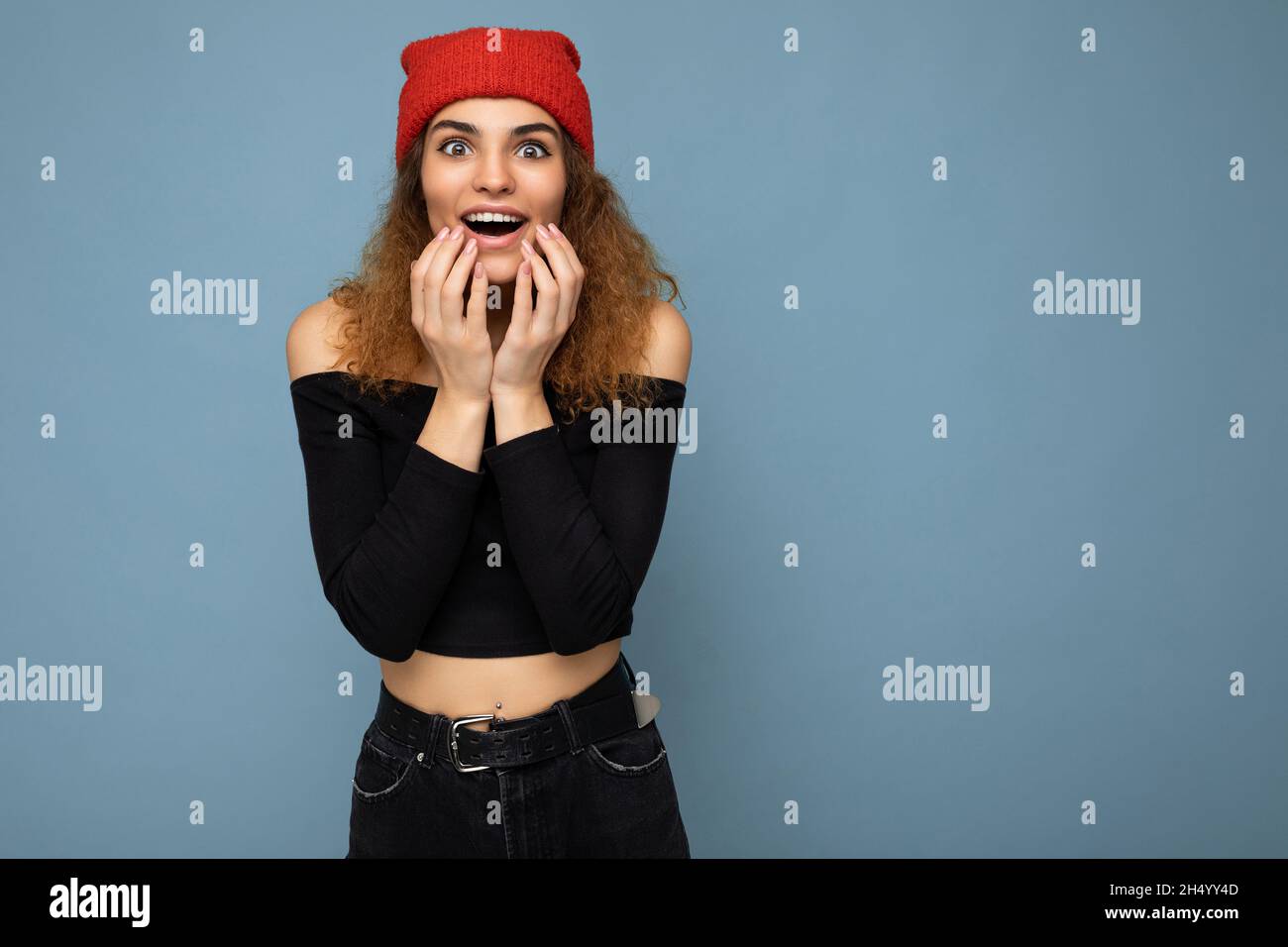 Photo de jeune émotif choqué étonné beau vin brunet femme ondulée-cheveux avec des émotions sincères portant noir crop top et chapeau rouge isolé sur bleu Banque D'Images