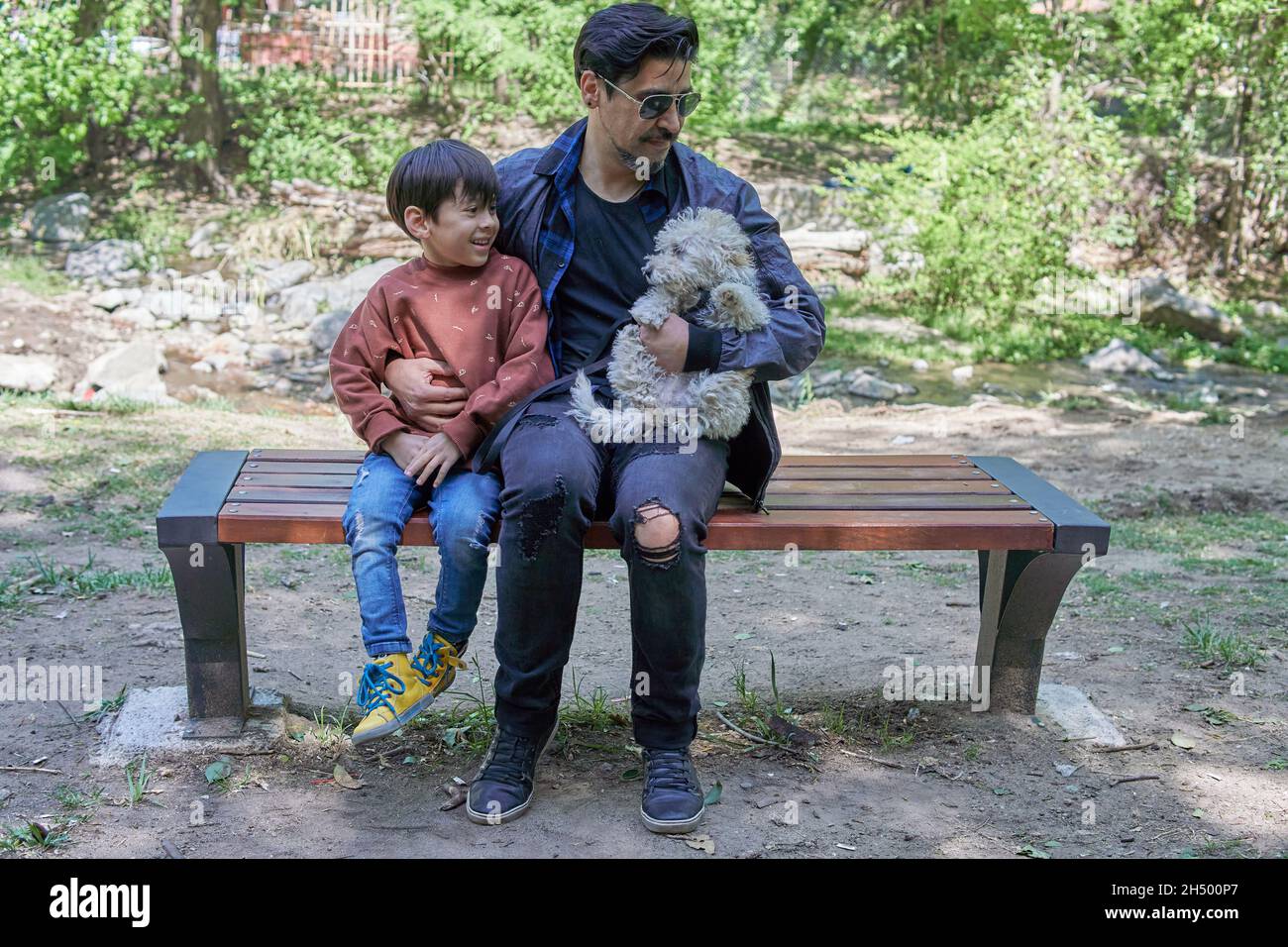 Père et fils latino-américains, et un chiot blanc en peluche, assis sur un banc de bois, reposant après une promenade le long de la crique. Horizontal.Gamme vac Banque D'Images