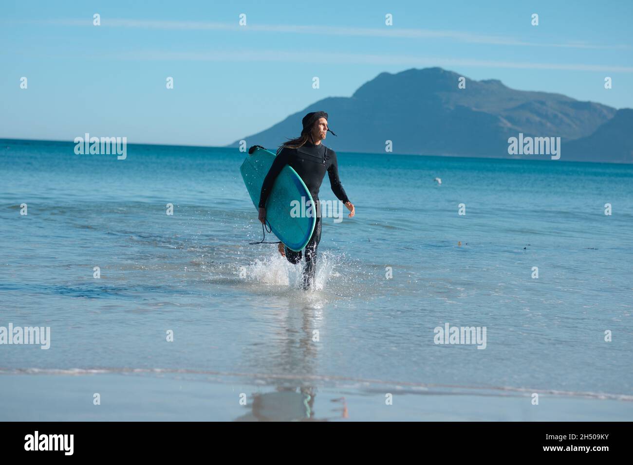 Homme avec planche de surf courant tout en barbotant de l'eau sur la plage pendant la journée ensoleillée Banque D'Images