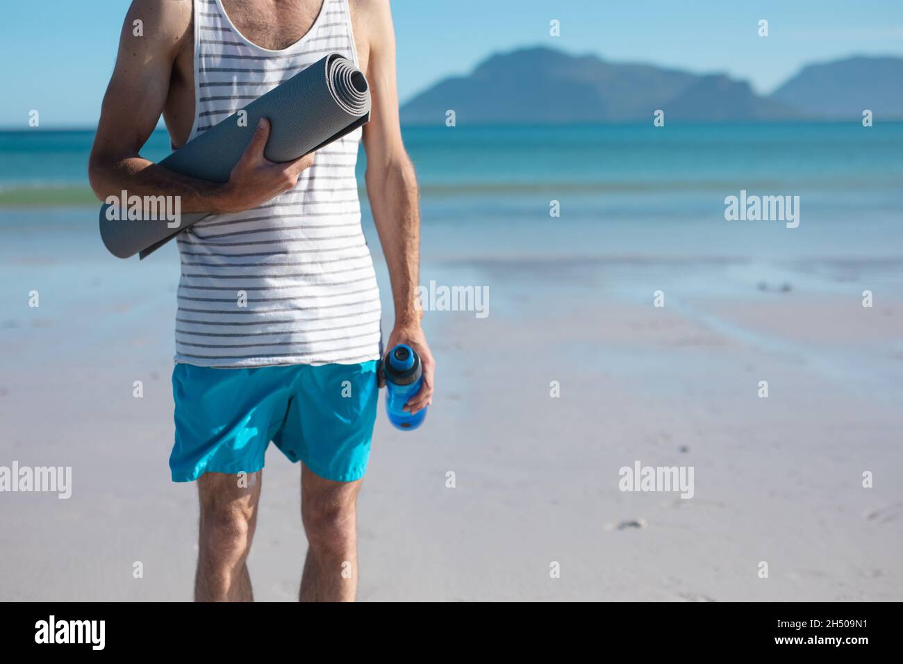 Section médiane de l'homme tenant un tapis de yoga et une bouteille d'eau à la plage le jour ensoleillé Banque D'Images