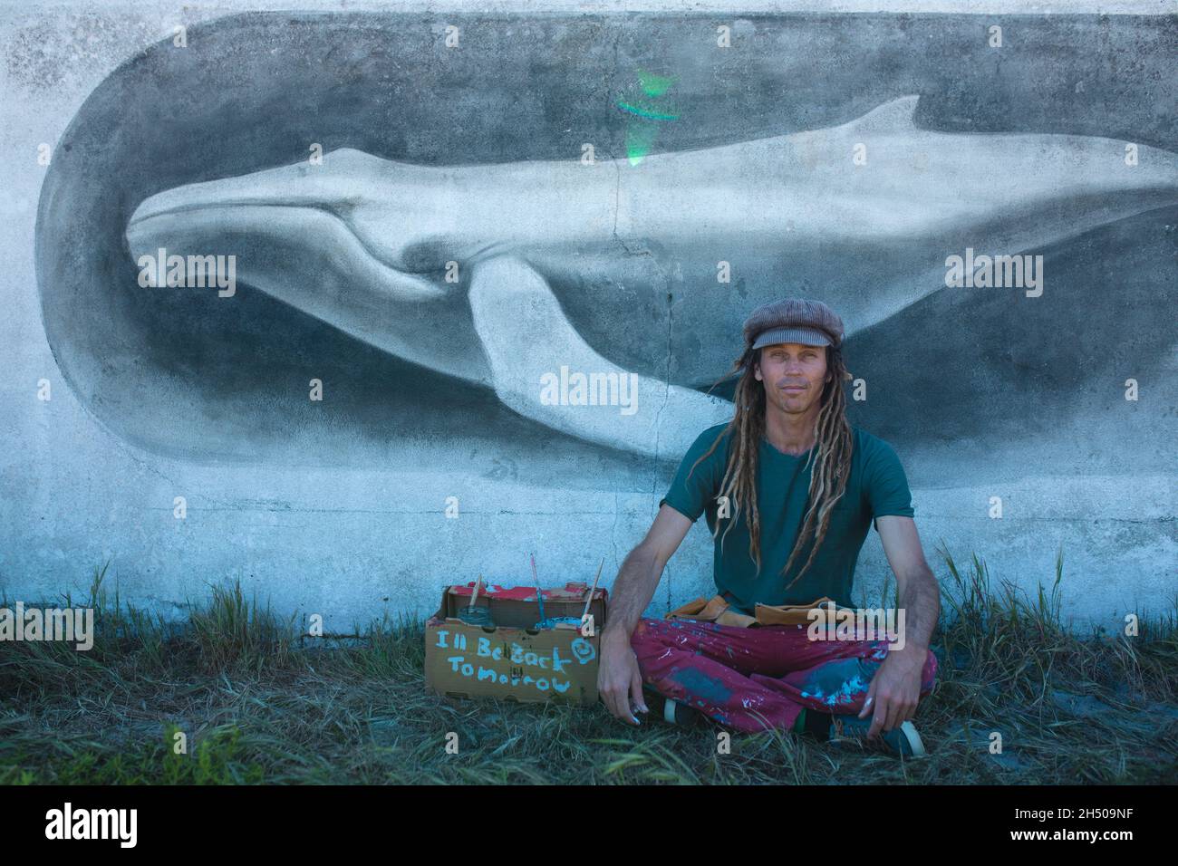 Fier homme artiste hippster assis à pattes croisées devant sa peinture murale de baleine sur le mur Banque D'Images