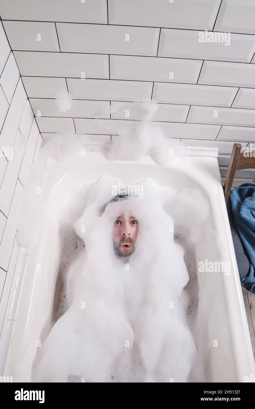 L'homme est baigné dans une baignoire, il est couvert de suds de savon luxuriants, seul son visage avec une expression drôle est visible. Banque D'Images
