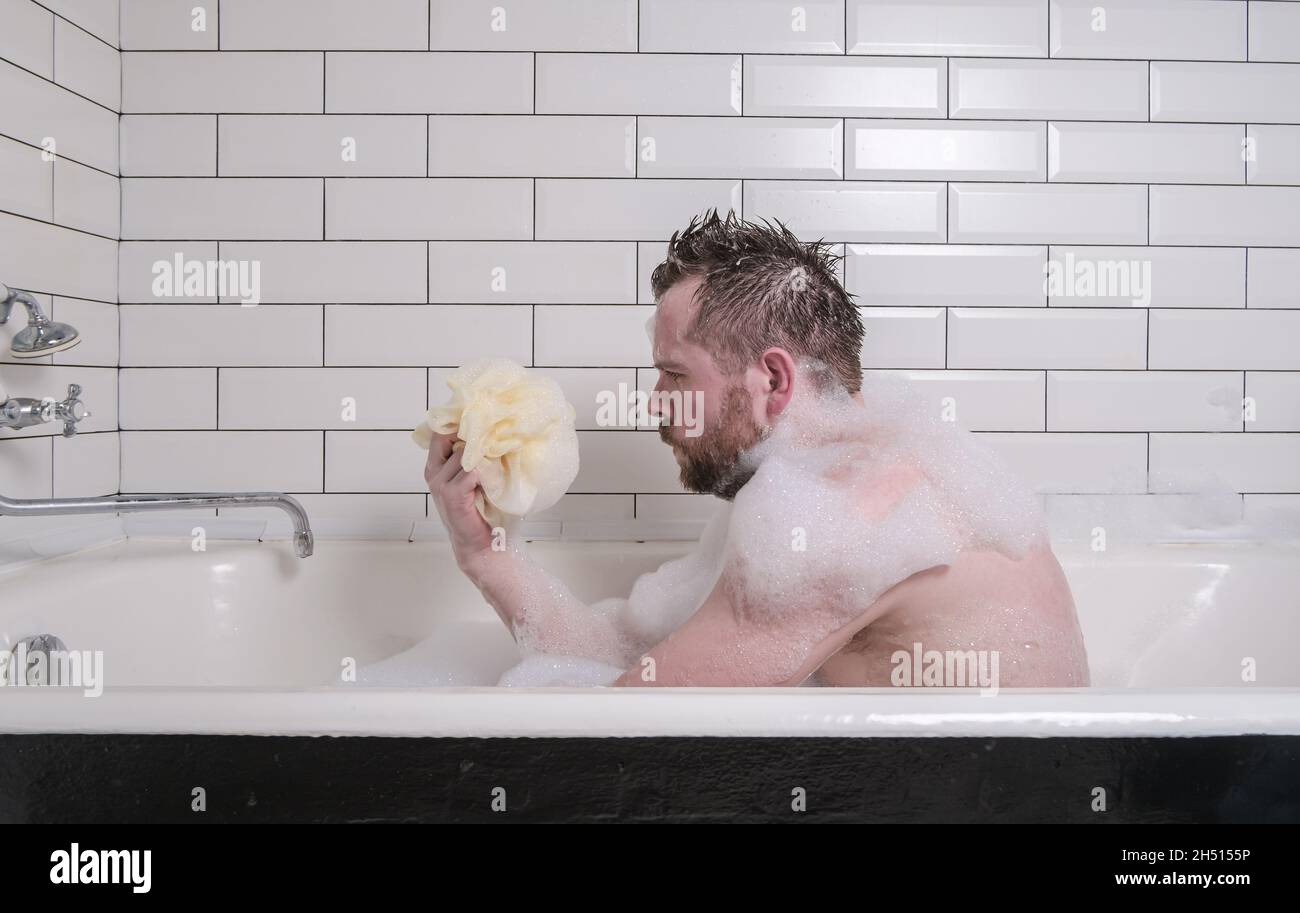 Triste homme baigné dans un bain avec des suds de savon luxuriants, il tient une éponge dans ses mains et regarde soigneusement. Banque D'Images