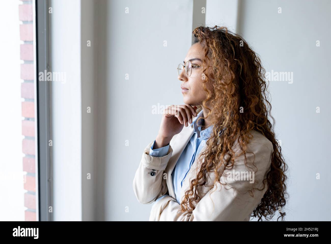 Femme d'affaires biraciale attentionnée regardant à travers la fenêtre tout en se tenant avec la main sur le menton dans le bureau Banque D'Images