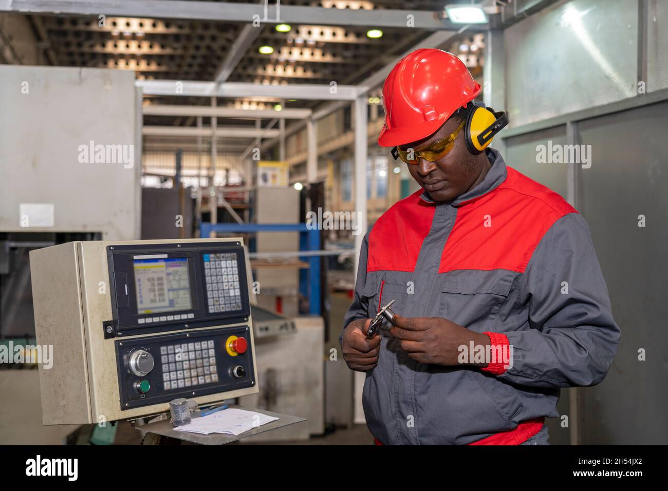 Portrait à la taille haute d'un ouvrier industriel noir en casque rouge, lunettes de sécurité jaunes, protecteurs d'oreilles et uniforme de travail à côté du contrôleur CNC. Banque D'Images