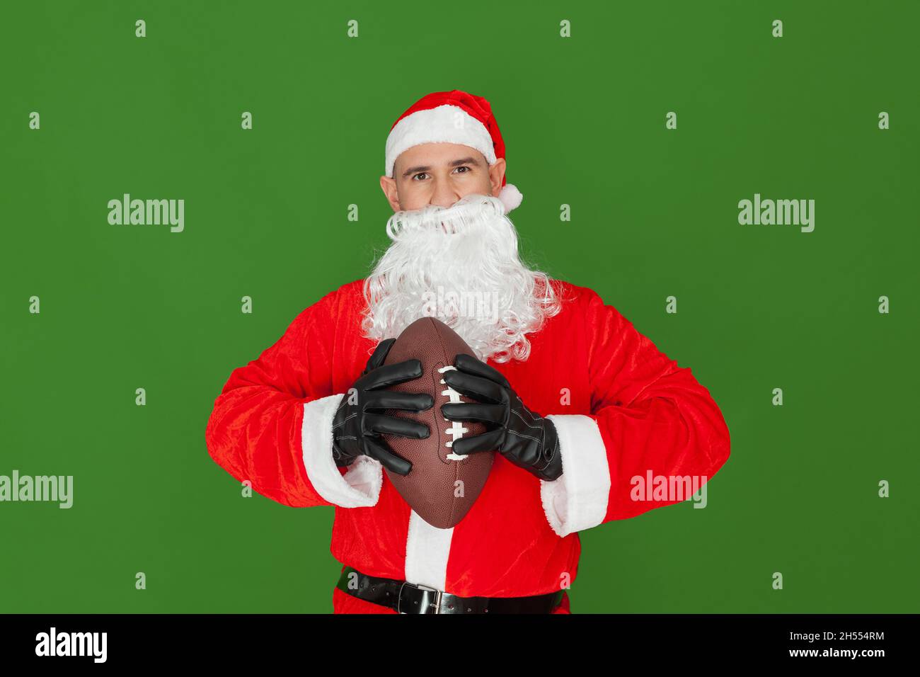 Un homme caucasien habillé comme le Père Noël regardant droit devant lui tenant une balle de football américain marron dans ses mains.L'arrière-plan est vert. Banque D'Images