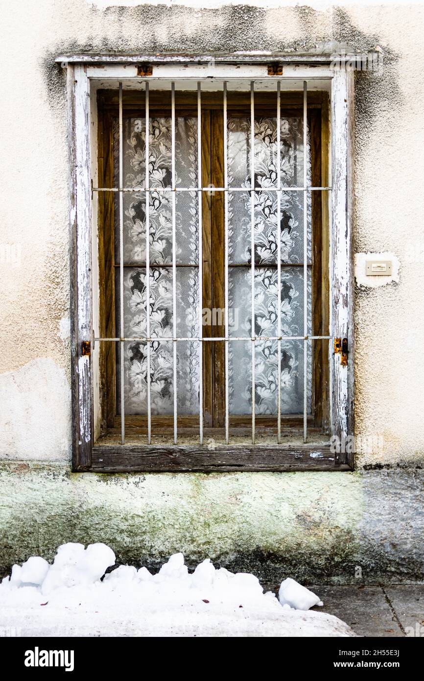 Ancienne fenêtre en bois avec barres et rideaux en fer, neige au sol Banque D'Images