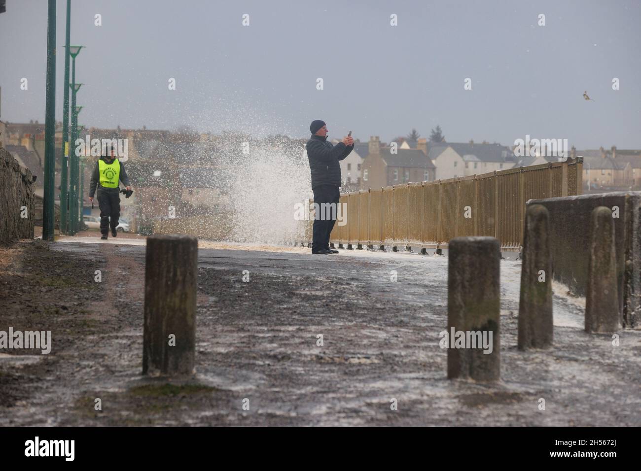 Thurso, Écosse.Novembre 7 2021.Un homme sur une promenade recouverte de mousse prend une photo tandis que des vents violents envoient des vagues s'écraser derrière lui. Banque D'Images