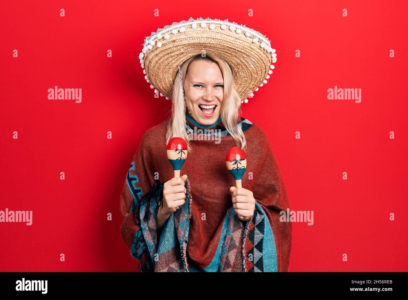 Belle femme blonde caucasienne portant un poncho mexicain festif et des maracas souriant et riant fort parce que drôle de blague folle. Banque D'Images