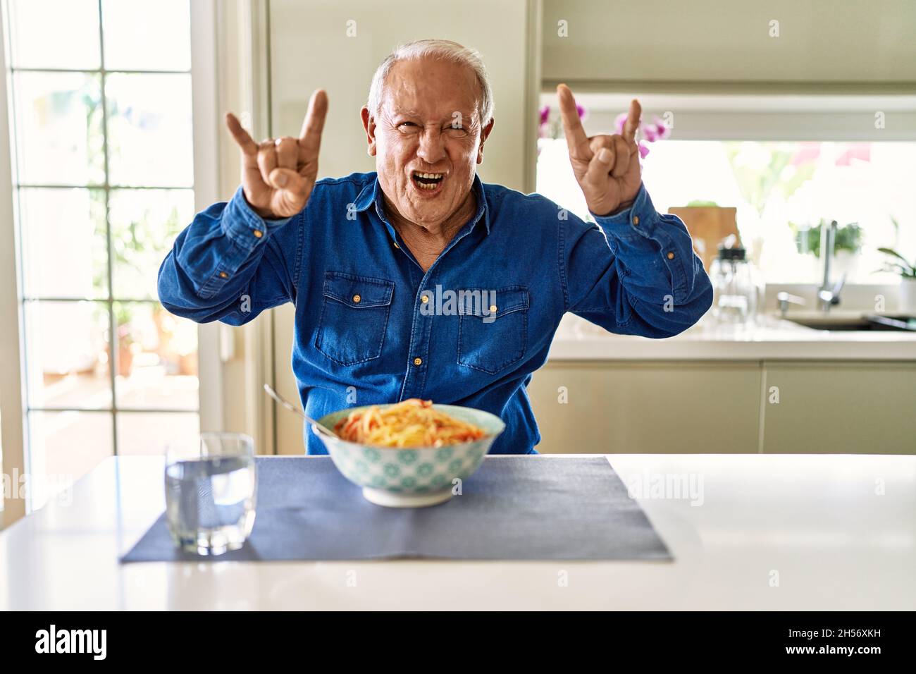 Homme âgé avec des cheveux gris mangeant des spaghetti de pâtes à la maison criant avec l'expression folle faisant le symbole de rock avec les mains vers le haut. Star de la musique. Concept lourd. Banque D'Images