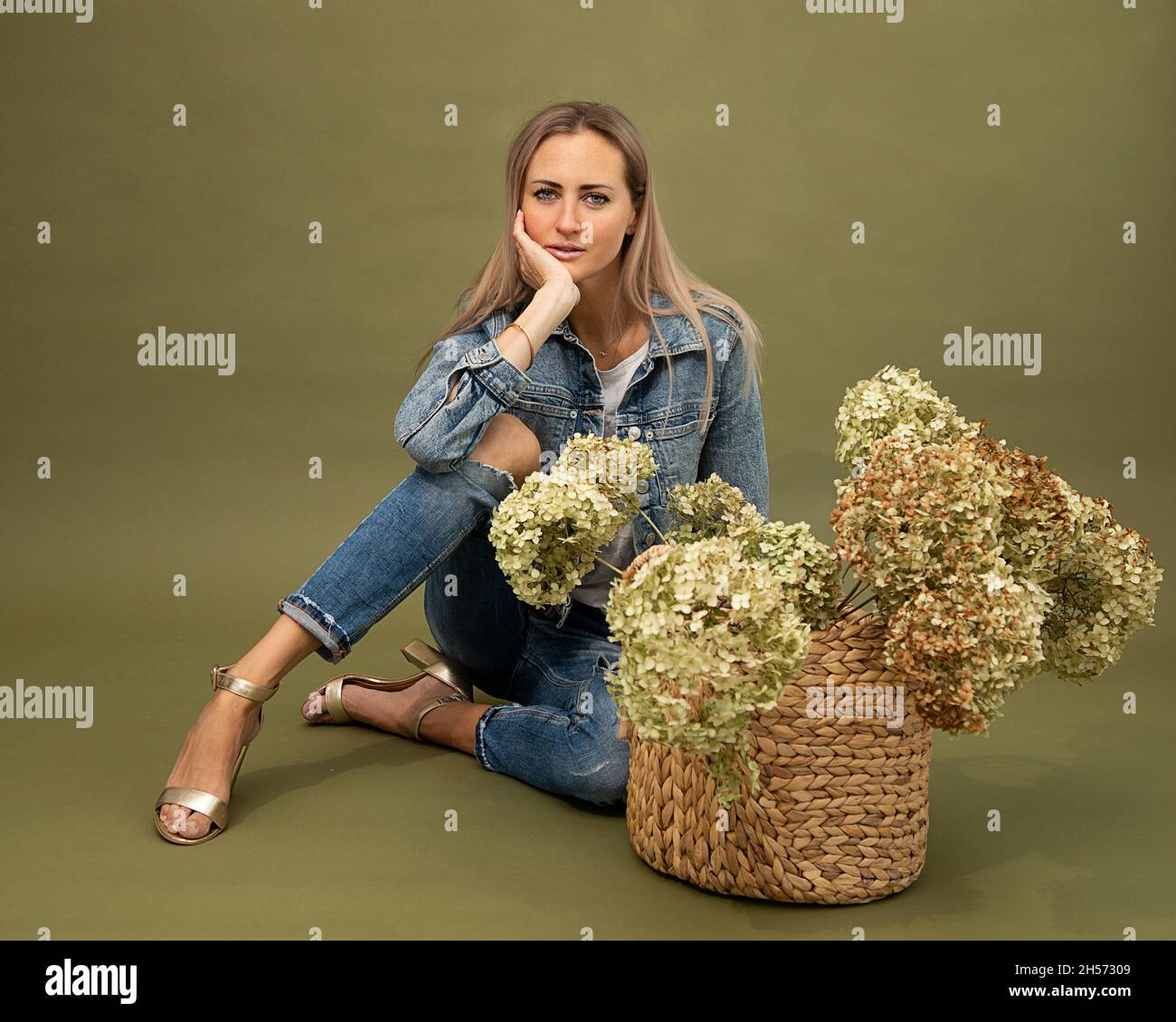 Portrait de modèle blond à la mode 30-35 ans fleuriste en denim posant sur fond d'olive en studio, fleurs d'hortensia séchées en buscrocket.Apparence Banque D'Images