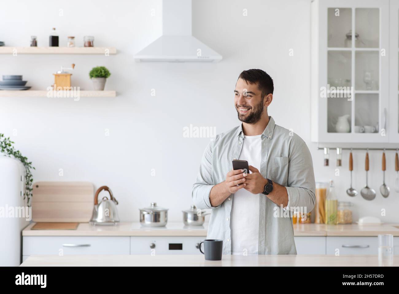 Bon homme de race blanche adulte avec un smartphone et une tasse de café regarder l'espace vide à l'intérieur de la cuisine Banque D'Images
