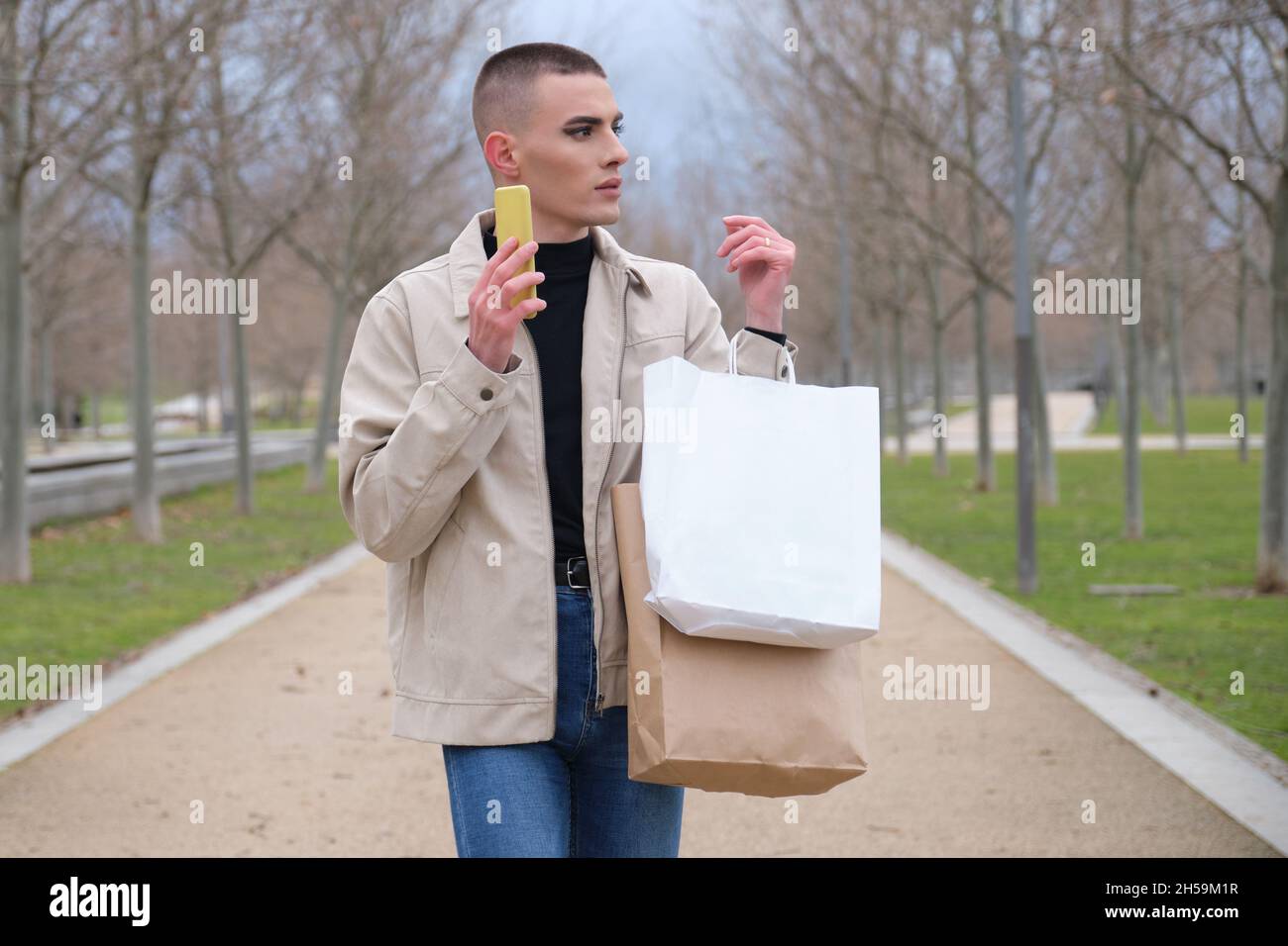 Jeune homme portant un maquillage tenant des sacs d'achats et un smartphone. Banque D'Images