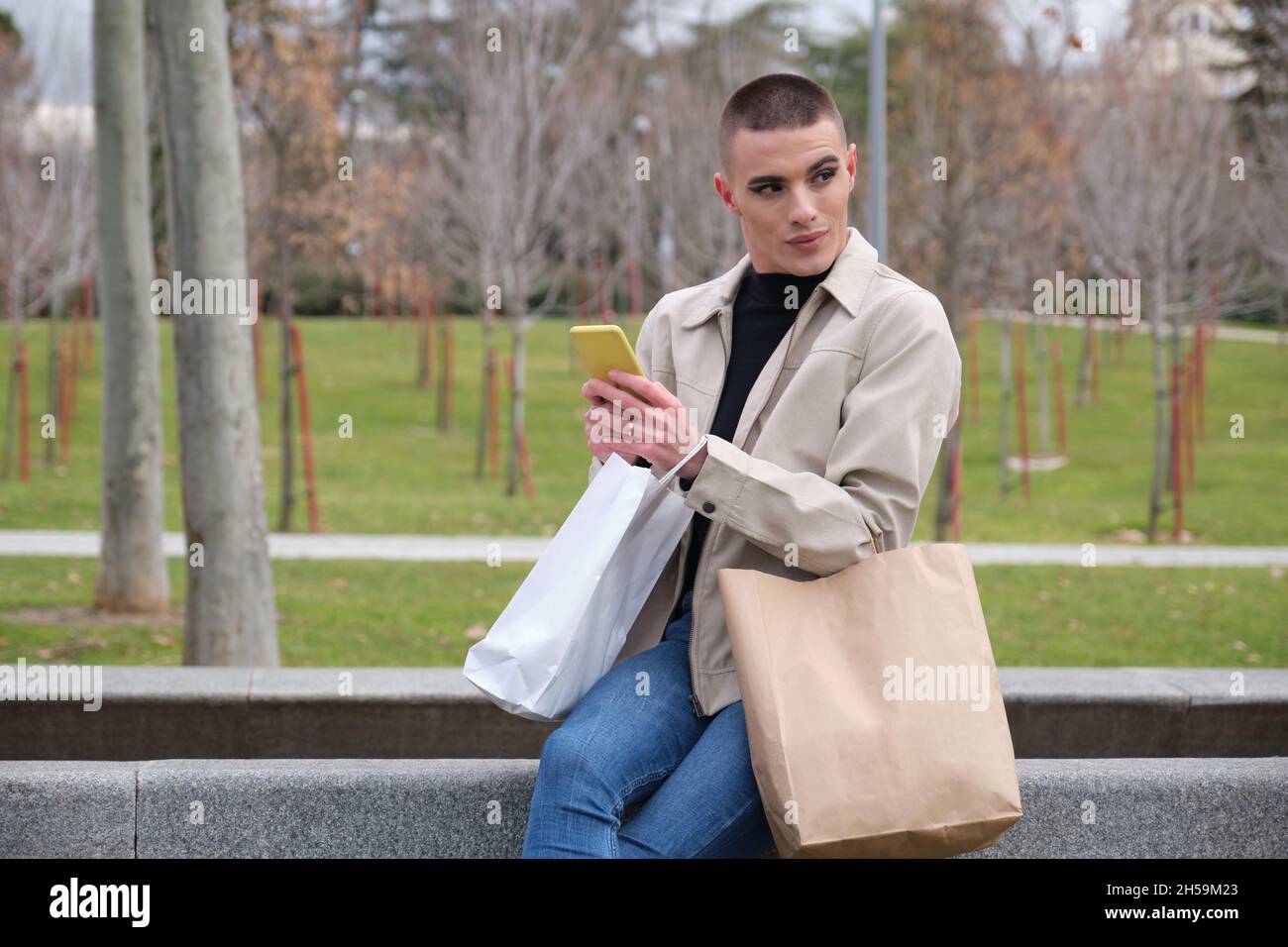 Jeune homme portant un maquillage tenant des sacs de shopping et bavardant sur un smartphone. Banque D'Images