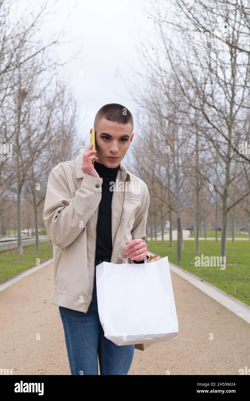 Jeune homme portant un maquillage tenant des sacs d'achats et parlant sur un smartphone. Banque D'Images