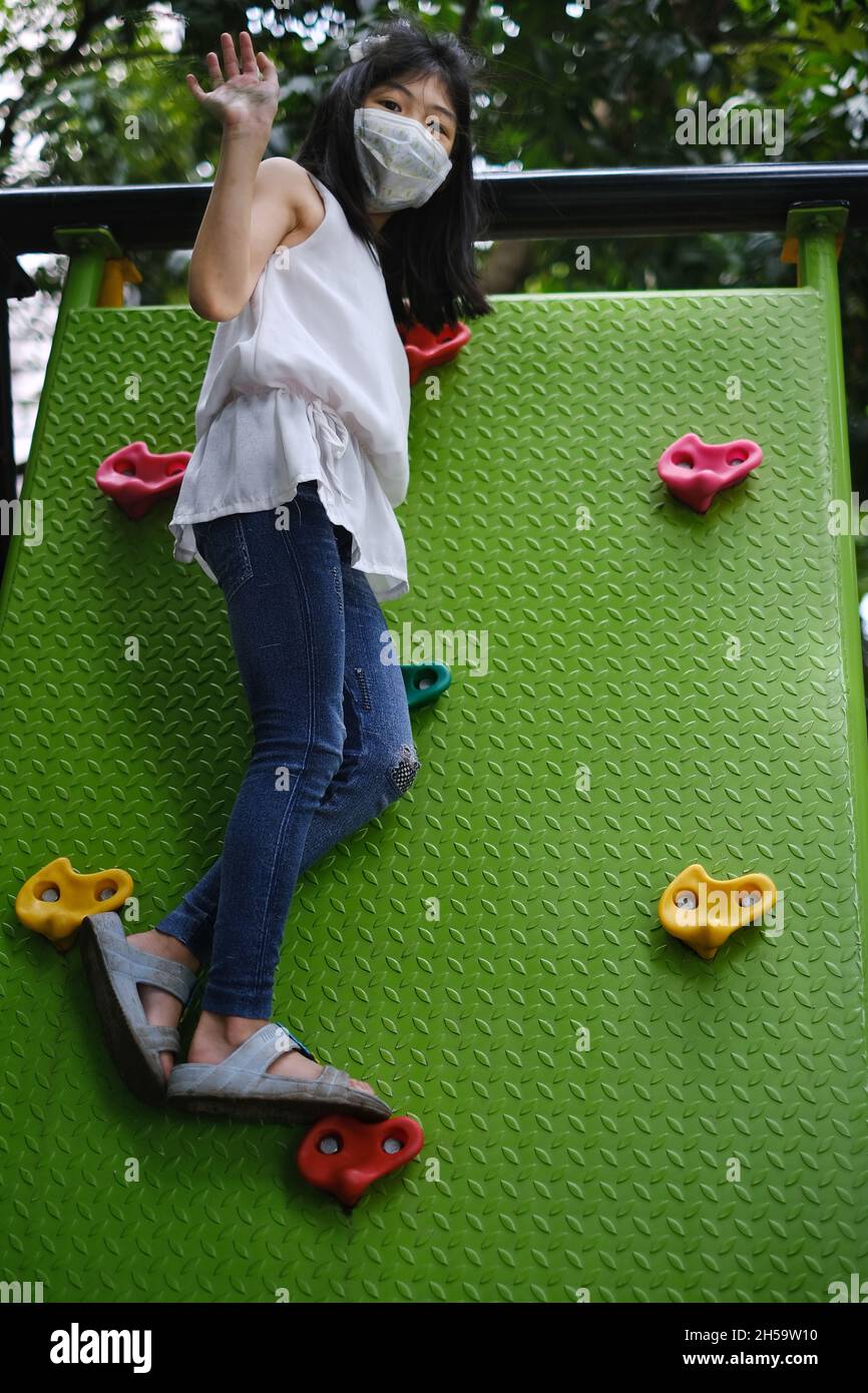 Une jolie jeune fille asiatique avec masque de visage joue seule dans le parc pendant la pandémie de Covid-19, grimpant une grande rampe verte sur le côté de la jungle gym. Banque D'Images