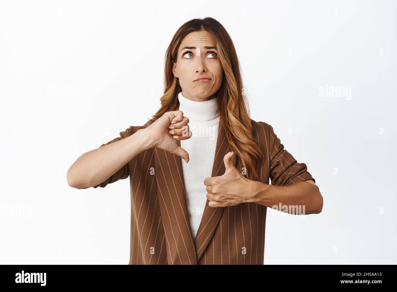 Indécis femme d'affaires d'âge moyen pesant la décision, montrant comme un geste déplaît, pouces vers le haut et pouces vers le bas, regardant vers le haut et pensant, blanc Banque D'Images