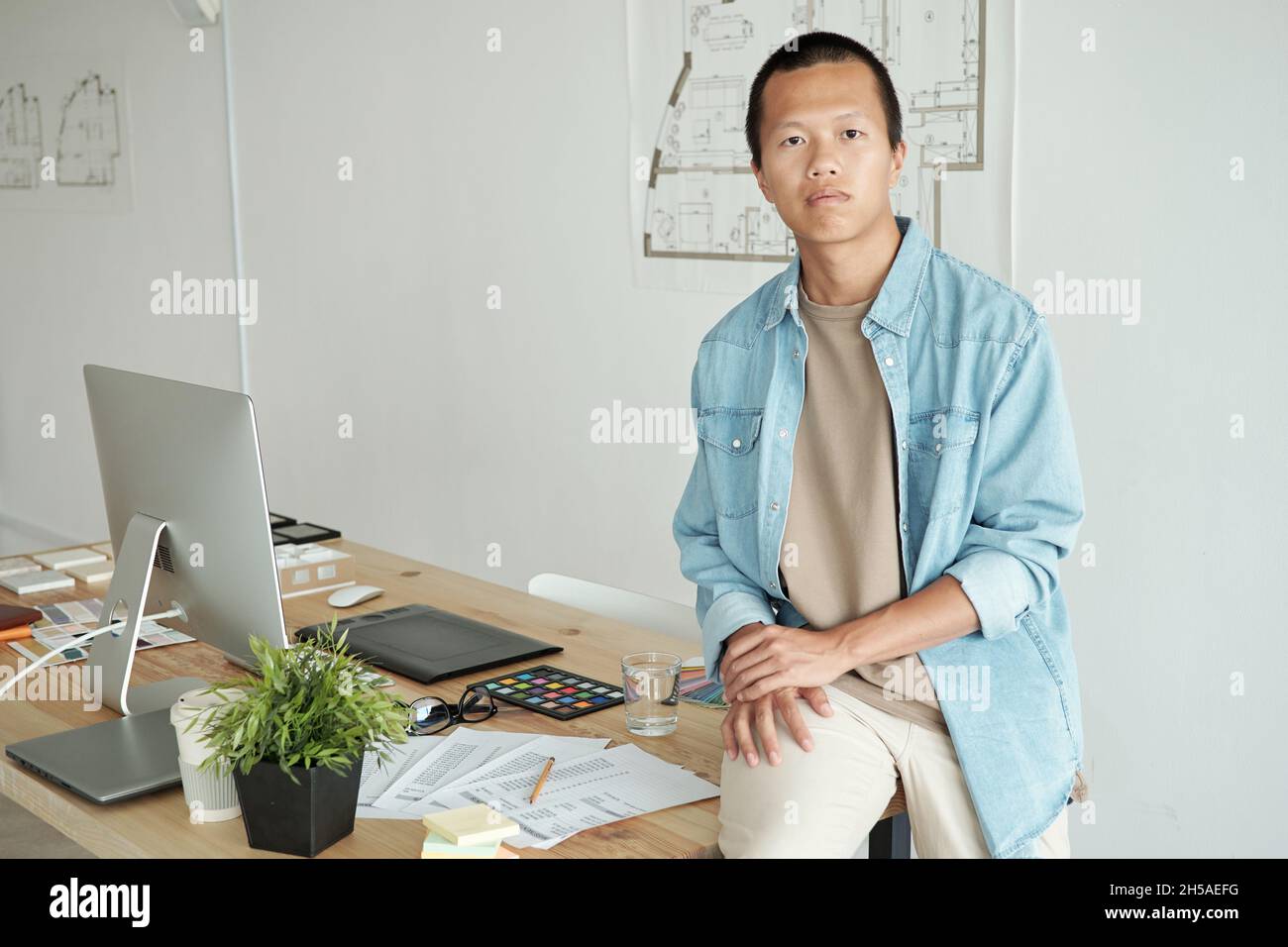 Jeune homme d'affaires chinois erious assis sur une table avec des papiers, une palette de couleurs, des échantillons, une tablette graphique et un écran d'ordinateur Banque D'Images