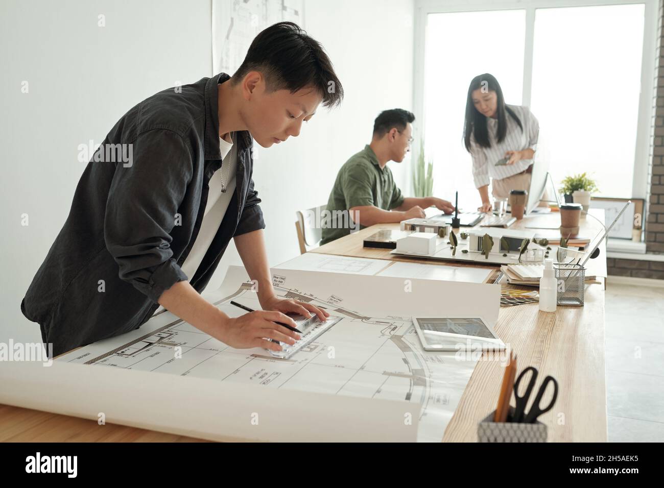 Jeune femme architecte ou ingénieur dessinant un croquis tout en se pliant sur la table contre ses deux collègues dans un grand bureau d'espace ouvert Banque D'Images