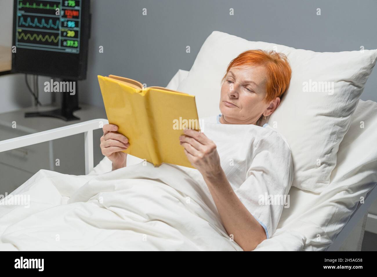 Femme âgée couchée au lit dans la salle d'hôpital et lisant un livre, elle se rétablissant après la maladie Banque D'Images
