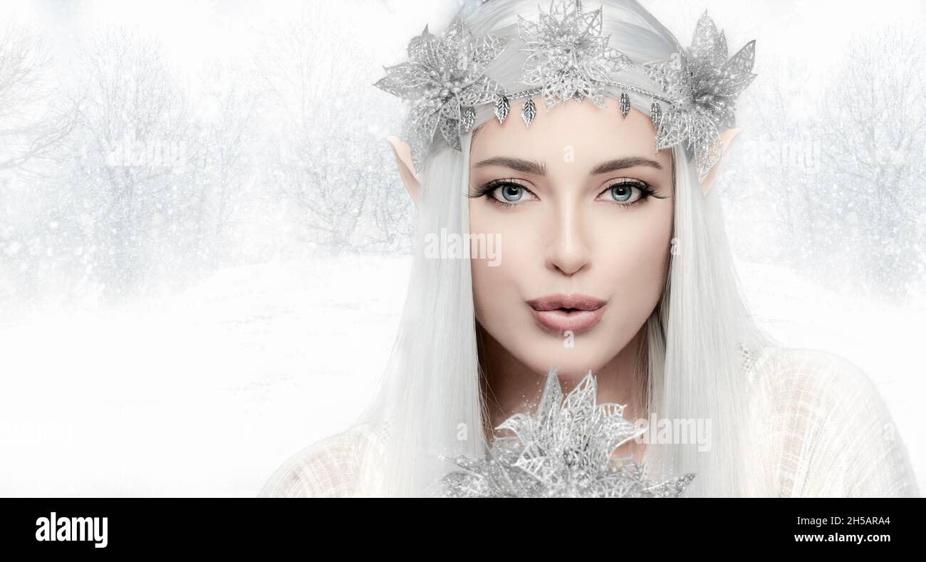 Belle jeune femme dans élégant costume de reine d'hiver soufflant de l'air froid sur la fleur fragile et regardant l'appareil photo sur fond de campagne d'hiver Banque D'Images