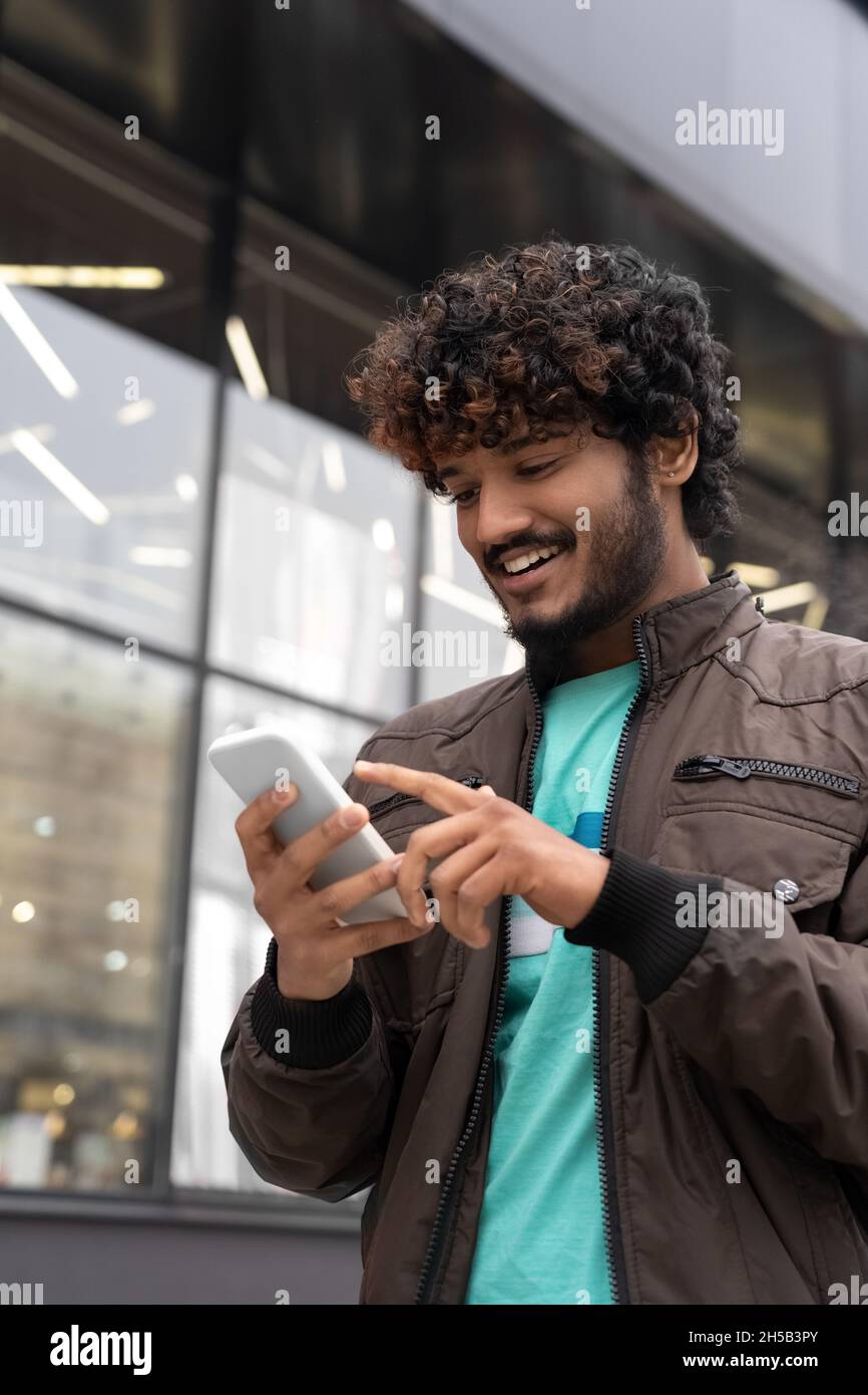 Joyeux souriant jeune Indien marchant dans la rue de la ville en utilisant le téléphone portable Banque D'Images