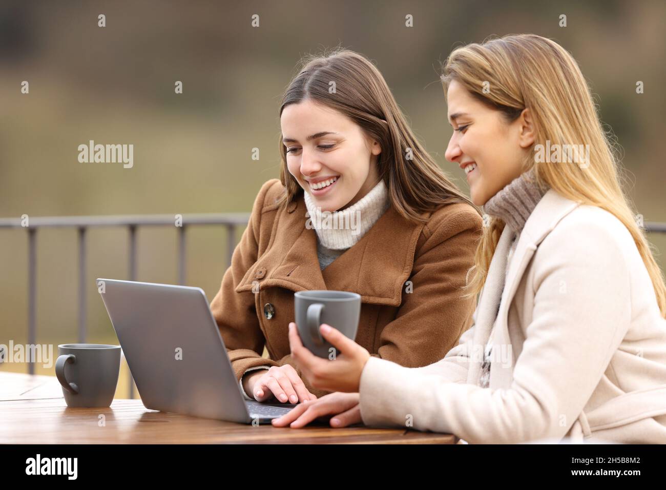 Deux heureux amis qui s'archivont sur leur ordinateur portable en hiver buvant un café assis sur une terrasse Banque D'Images