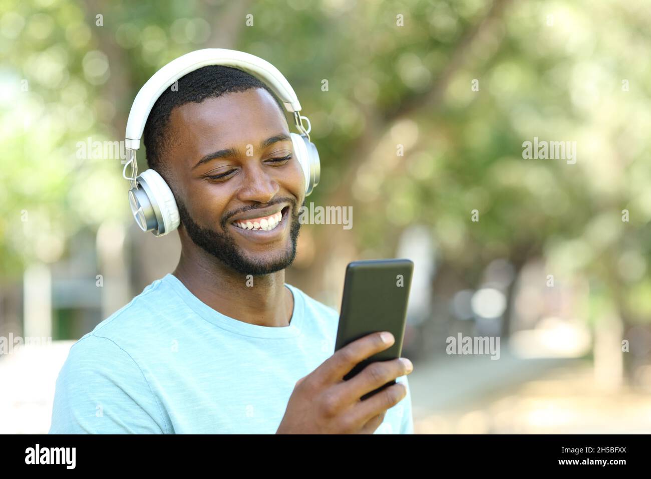Homme heureux avec peau noire écoutant de la musique avec des écouteurs et un smartphone marchant dans un parc Banque D'Images