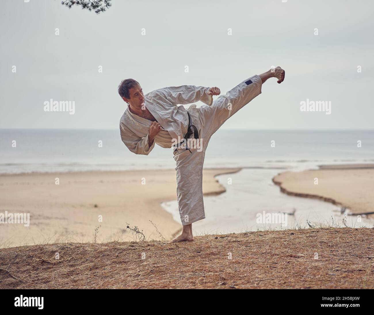 Carate homme dans un vieux kimono et ceinture noire de l'entraînement de  coup de pied côté à la mer.Concept des arts martiaux.La rivière coule dans  la mer à l'arrière-plan Photo Stock -
