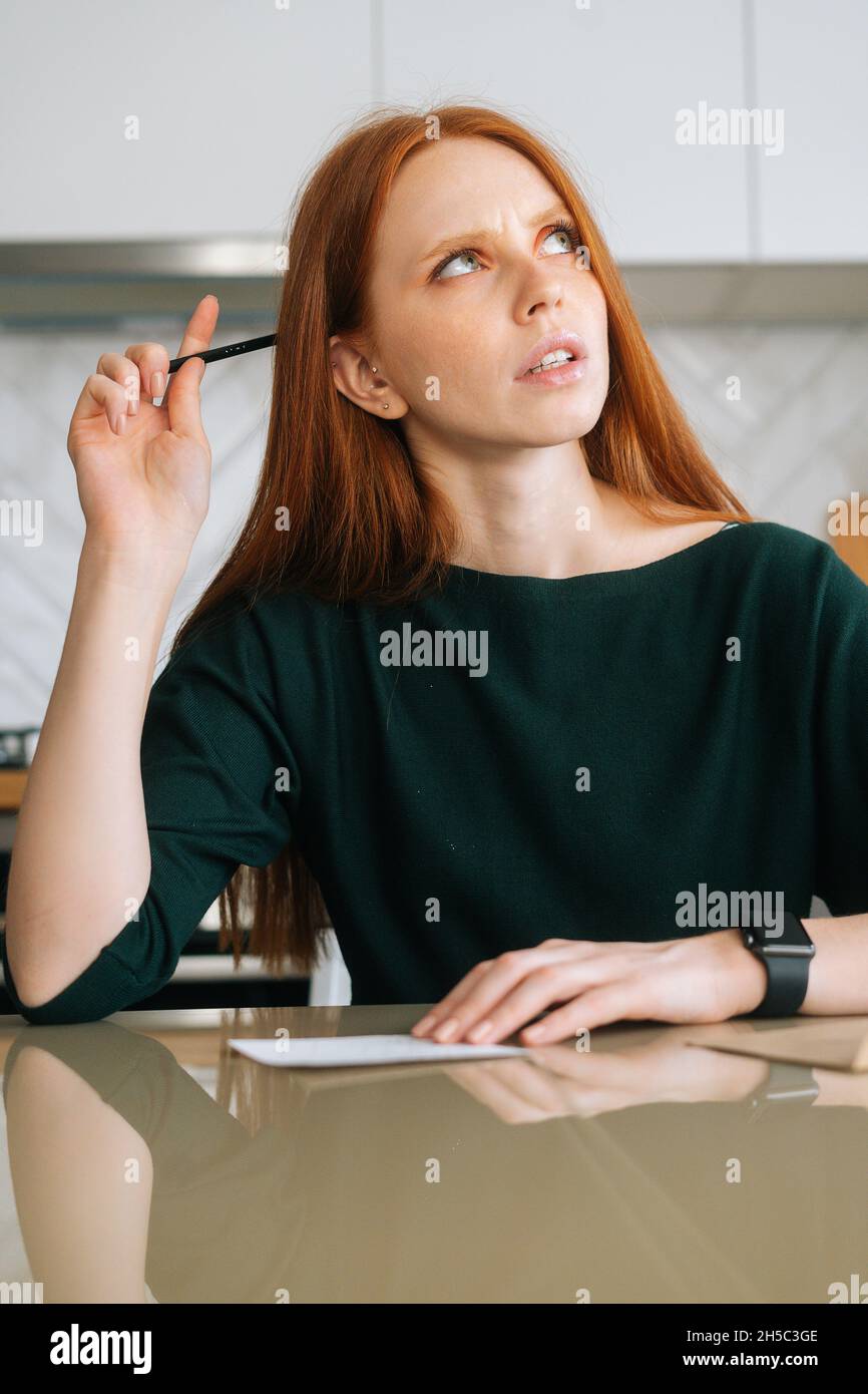 Photo verticale d'une jeune femme en train de réfléchir écrivant une lettre manuscrite assise à une table avec une enveloppe dans la cuisine avec intérieur lumineux, en regardant vers le haut. Banque D'Images