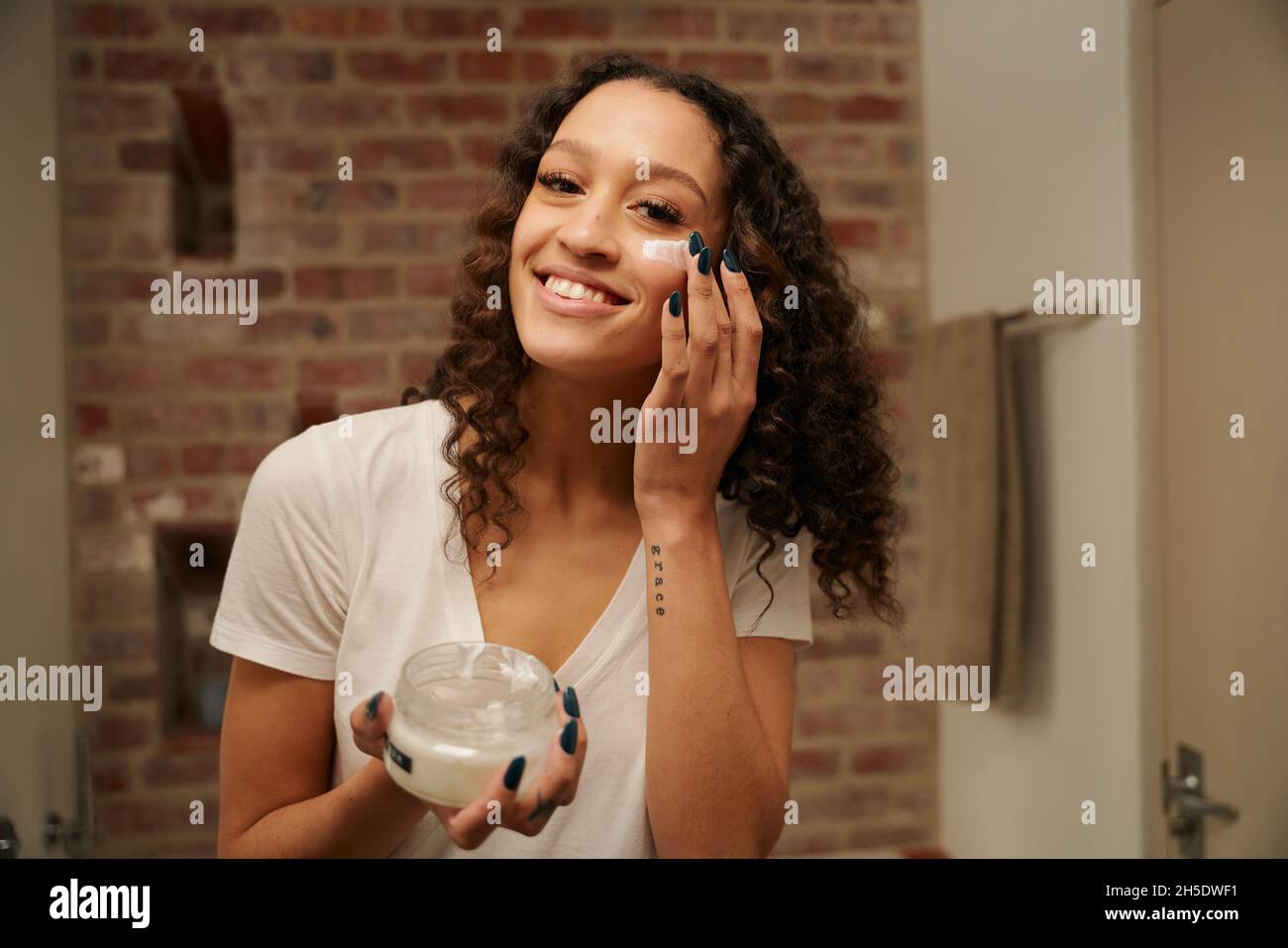 Multi-culturel jeune adulte femme smilimg tout en appliquant hydratant sur son visage dans son miroir de salle de bains Banque D'Images