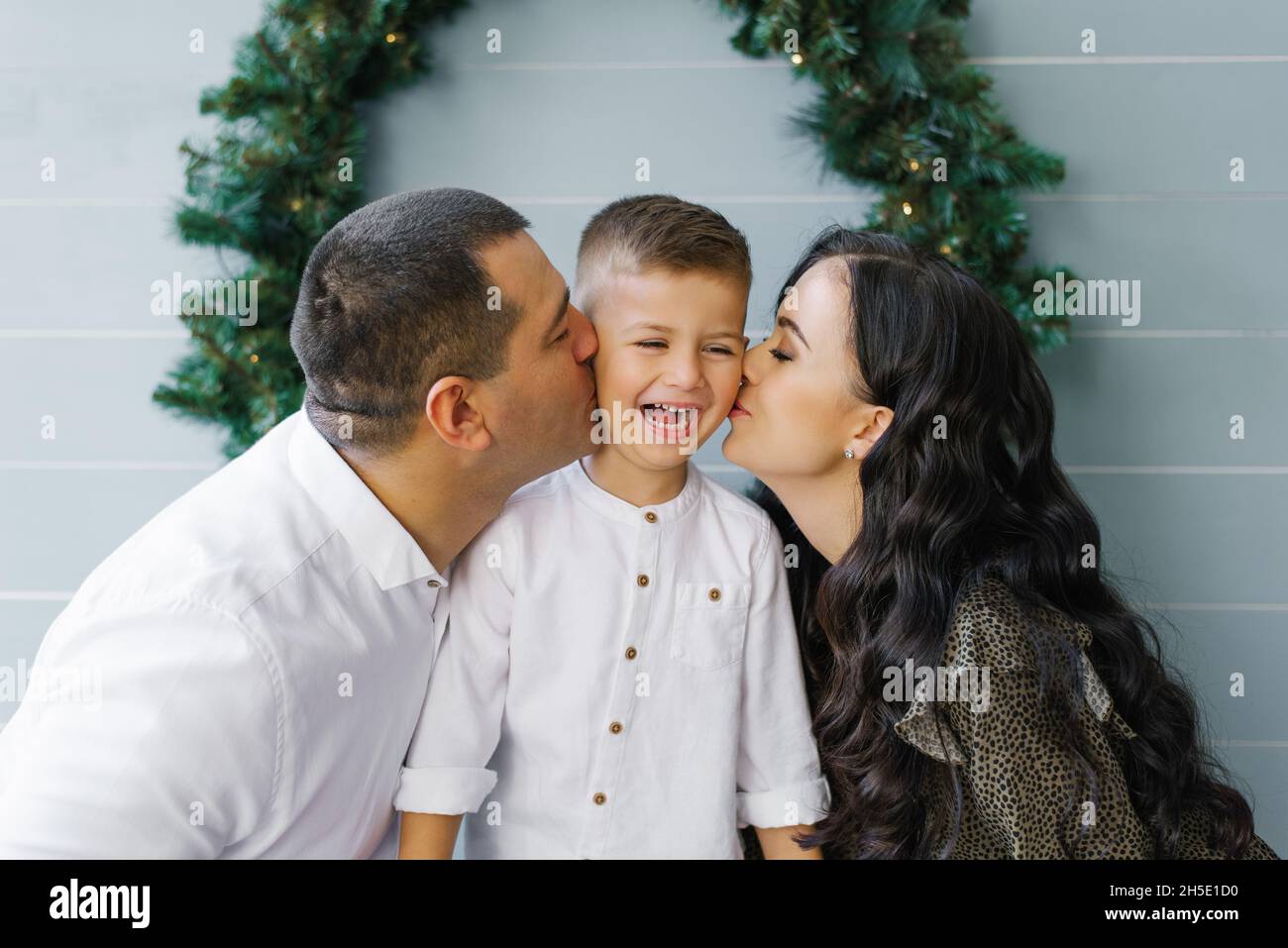 Belle jeune maman et papa embrasser un petit fils mignon debout sur le fond d'une couronne de Noël sur le mur Banque D'Images