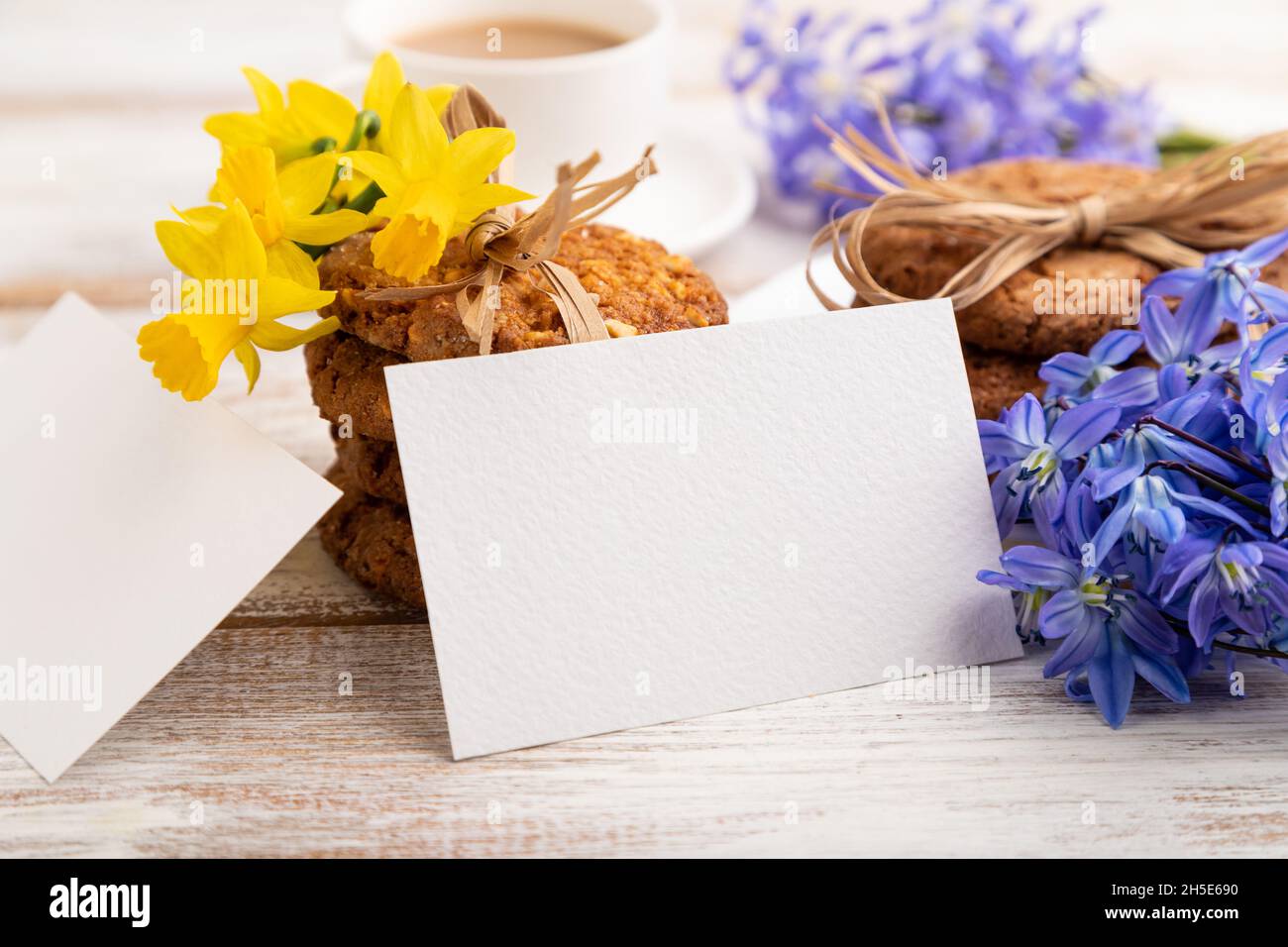 Carte de visite en papier blanc avec biscuits aux flocons d'avoine, cloches de fleurs de neige printanières, narcisses et tasse de café sur fond de bois blanc. Banque D'Images
