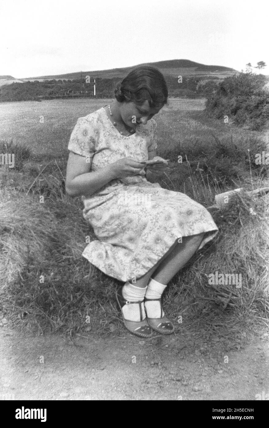 Années 1940, historique, une adolescente en robe d'été, des chaussettes de cheville et des chaussures à talons de sable, assis sur une banque herbeuse lisant un journal ou un petit livre, Angleterre, Royaume-Uni. Banque D'Images