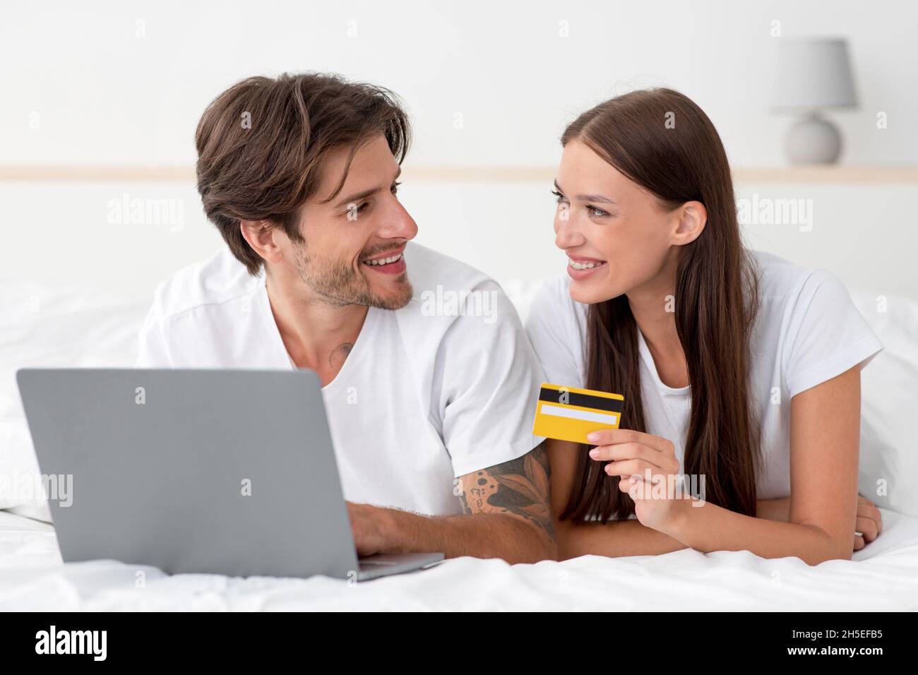 Une jeune femme et un mari européens joyeux se trouvent au lit, en payant l'achat et la commande Banque D'Images