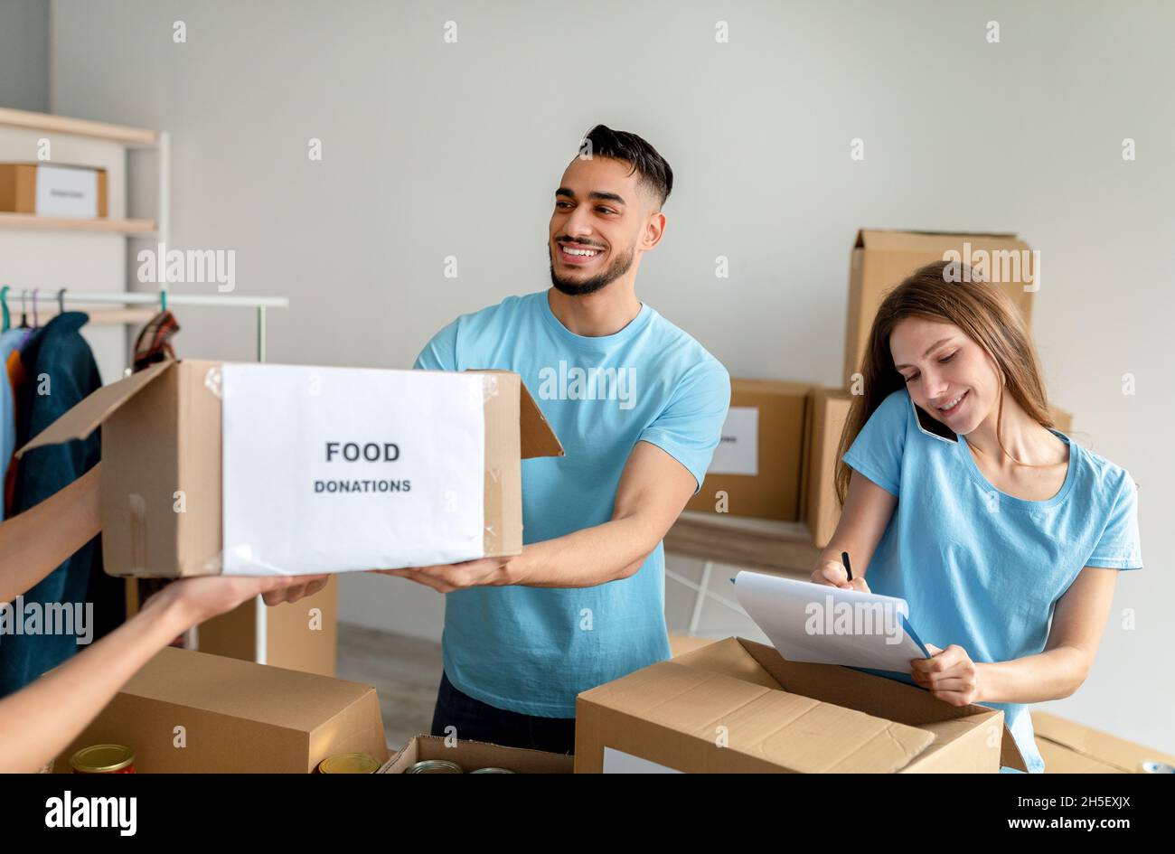 Des bénévoles heureux triant et emballant des dons alimentaires dans des boîtes en carton, une femme écrivant dans un presse-papiers Banque D'Images