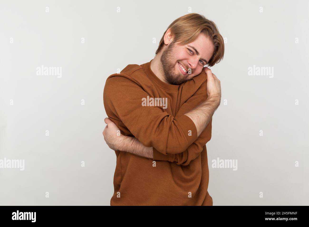 Portrait de l'homme beau heureux avec barbe portant un sweat-shirt, debout et s'embrassant avec le sourire crasseux appréciant, estime de soi positive.Prise de vue en studio isolée sur fond gris. Banque D'Images