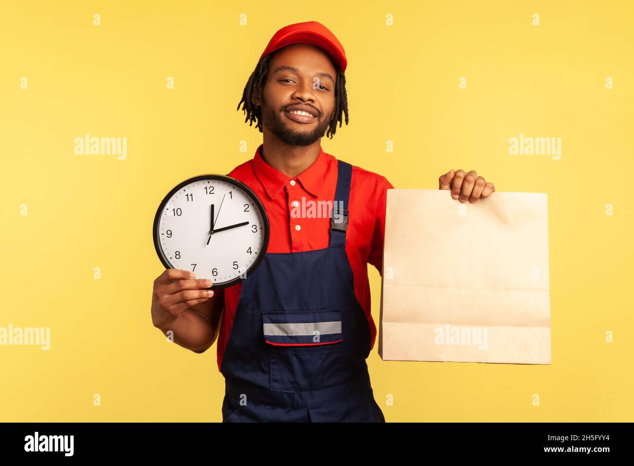 Livraison de nourriture, livraison à temps.Messager portant une combinaison et un T-shirt tenant une horloge et un colis, transport express par messagerie, services postaux.Studio d'intérieur isolé sur fond jaune Banque D'Images