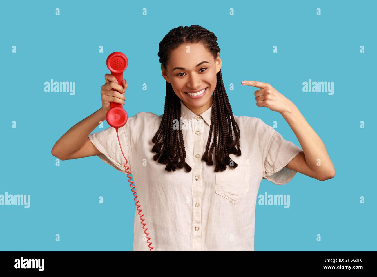 Portrait d'une femme souriante satisfaite avec des dreadlocks noirs tenant le téléphone rouge rétro, centre d'appels. Portant une chemise blanche.Studio d'intérieur isolé sur fond bleu. Banque D'Images
